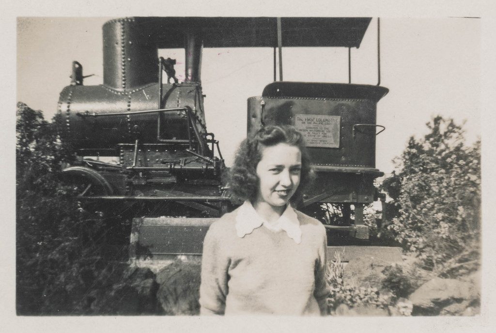 The Oregon Pony, first locomotive in the Pacific Northwest, sits unrestored with a young woma posing in the foreground. 