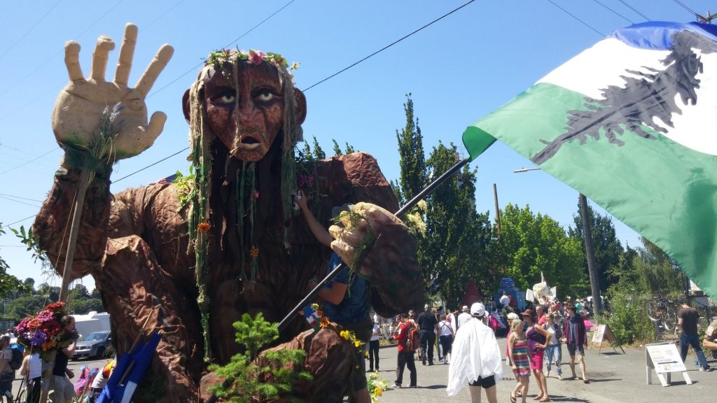 A giant Sasquatch puppet with a flower crown raises one hand at a street parade while the blue, white, and green Doug flag, marked with a black Douglas fir, waves in the foreground.